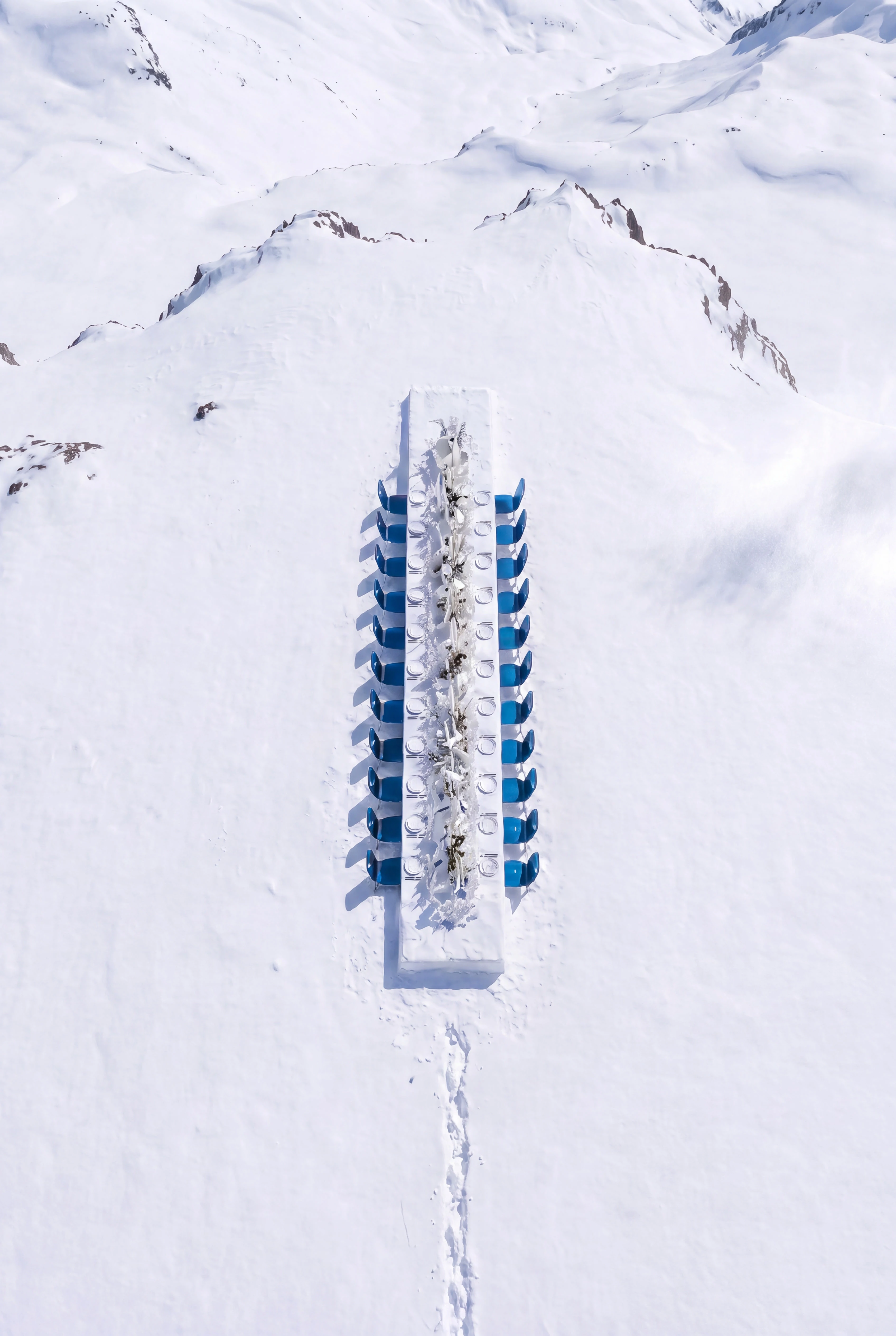 Vertical aerial view of a precisely aligned dining table and blue chairs in untouched snow — Spatial Design by Parallel Creative Agency — Uncharted Table, hospitality
